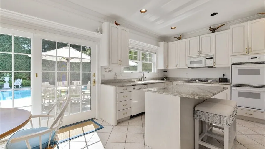 Una cocina luminosa con gabinetes blancos, una isla central y ventanas con vista a una piscina.