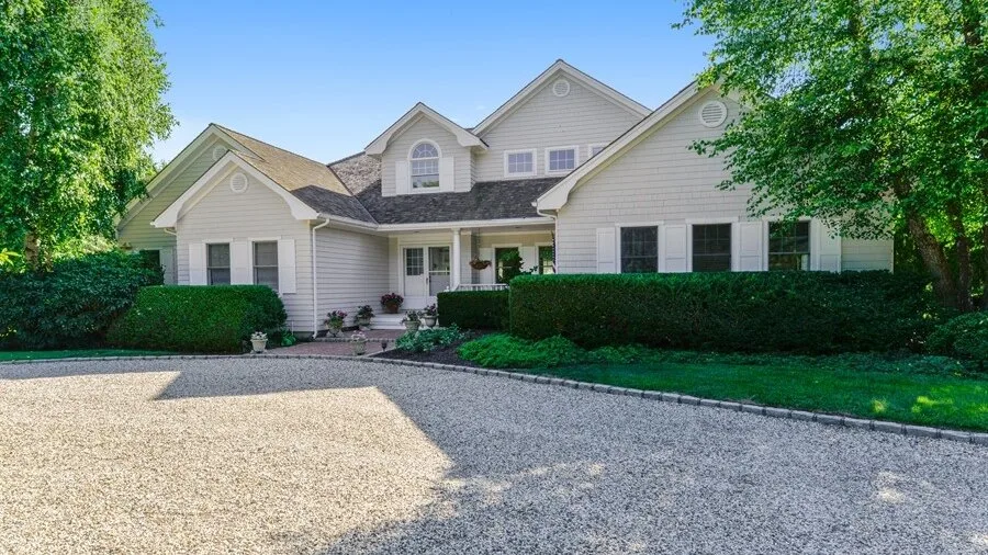 A large light-colored house stands behind a gravel driveway and green landscaping.