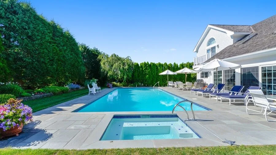 An outdoor pool and spa beside a large house, with lounge seating on the patio.