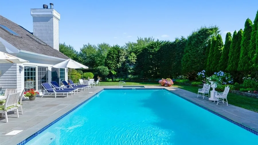 Una piscina al aire libre junto a un patio pavimentado y vegetación ornamental.