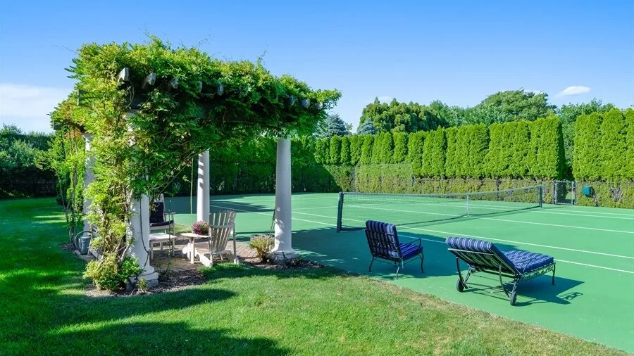 An outdoor tennis court with a net, seating, and a shaded garden pergola.