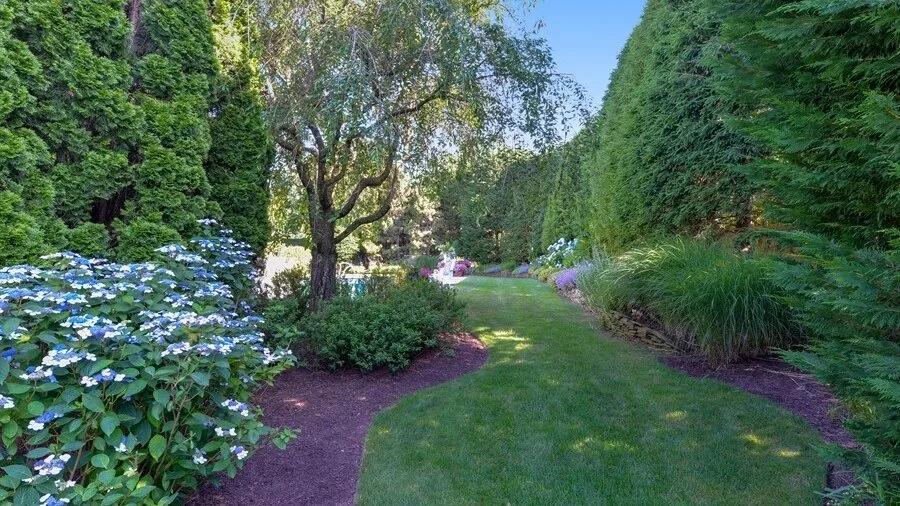 Un sendero de jardín pasa entre plantas con flores, arbustos y setos altos.