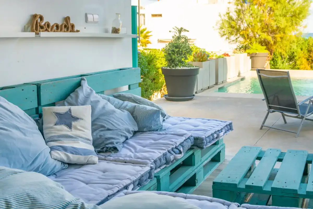 Una zona de estar exterior turquesa junto a una piscina, con cojines y plantas en macetas.