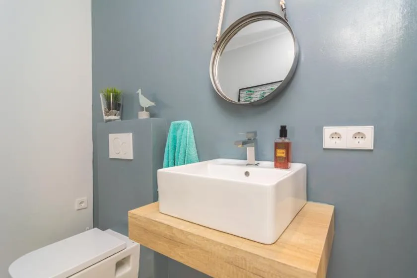 Un baño con un lavabo blanco sobre un mueble de madera y un espejo redondo.