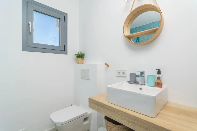 Un baño luminoso con un lavabo sobre un mueble de tono madera y un espejo redondo.