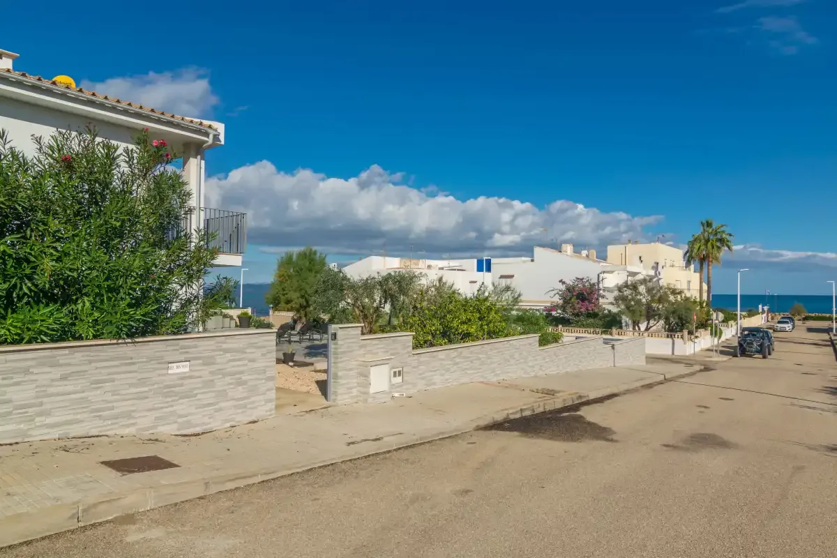 Una calle costera soleada con muros bajos, casas y el mar al fondo.