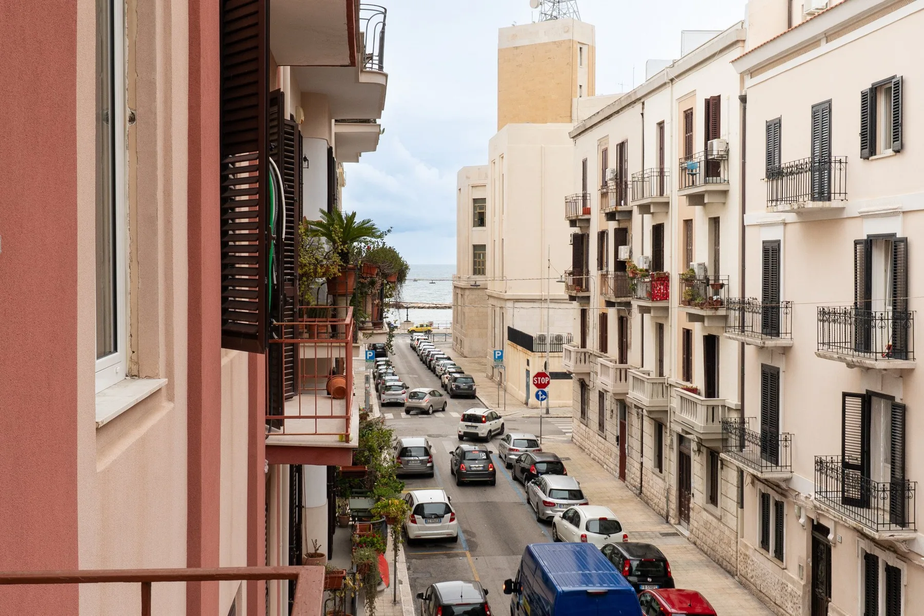 A narrow street lined with apartment buildings and parked cars, with the sea visible in the distance.