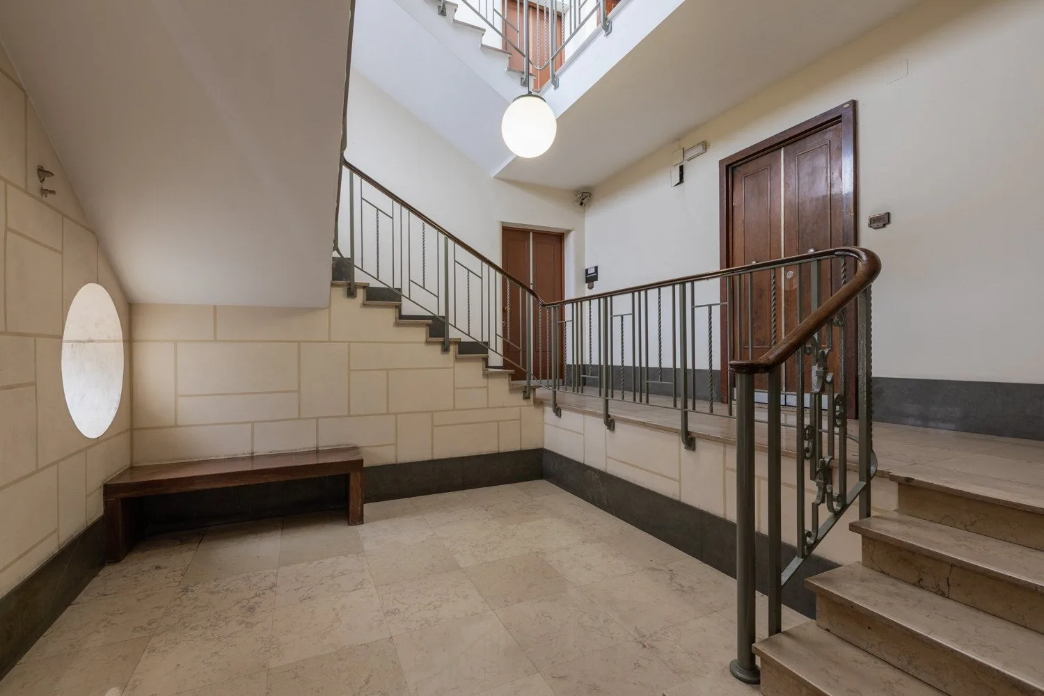 Una escalera interior con paredes de azulejos, barandillas oscuras y una lámpara colgante.