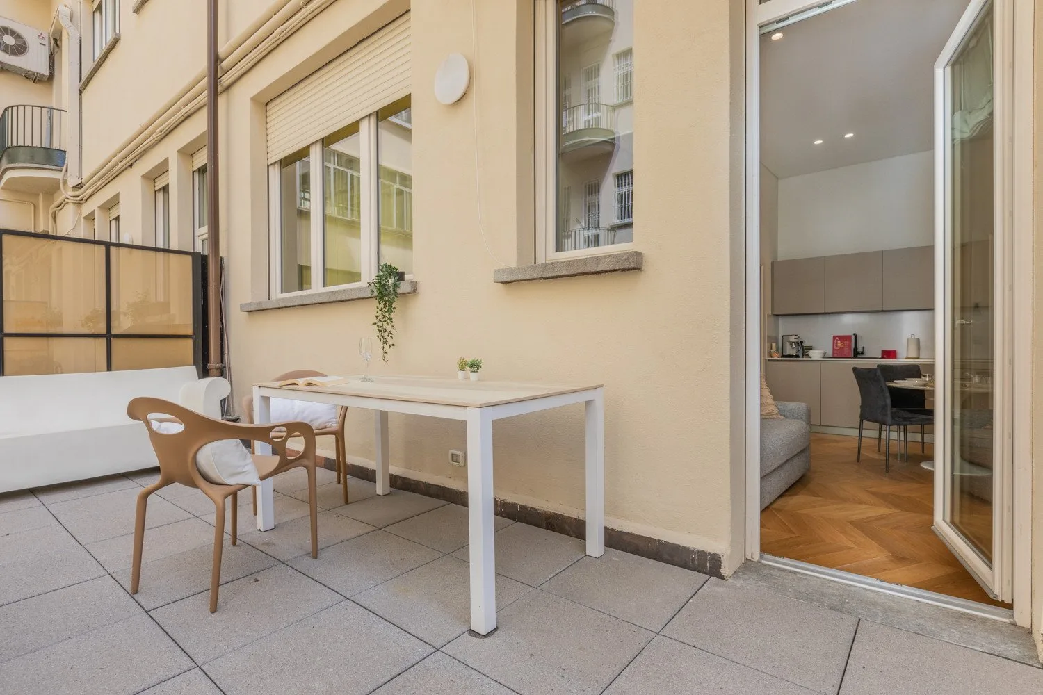 Una terraza pavimentada con una mesa pequeña y sillas junto a una puerta abierta.