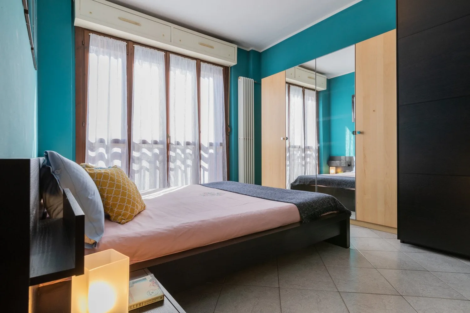 Un dormitorio con una cama junto a ventanas altas con cortinas y paredes color turquesa.