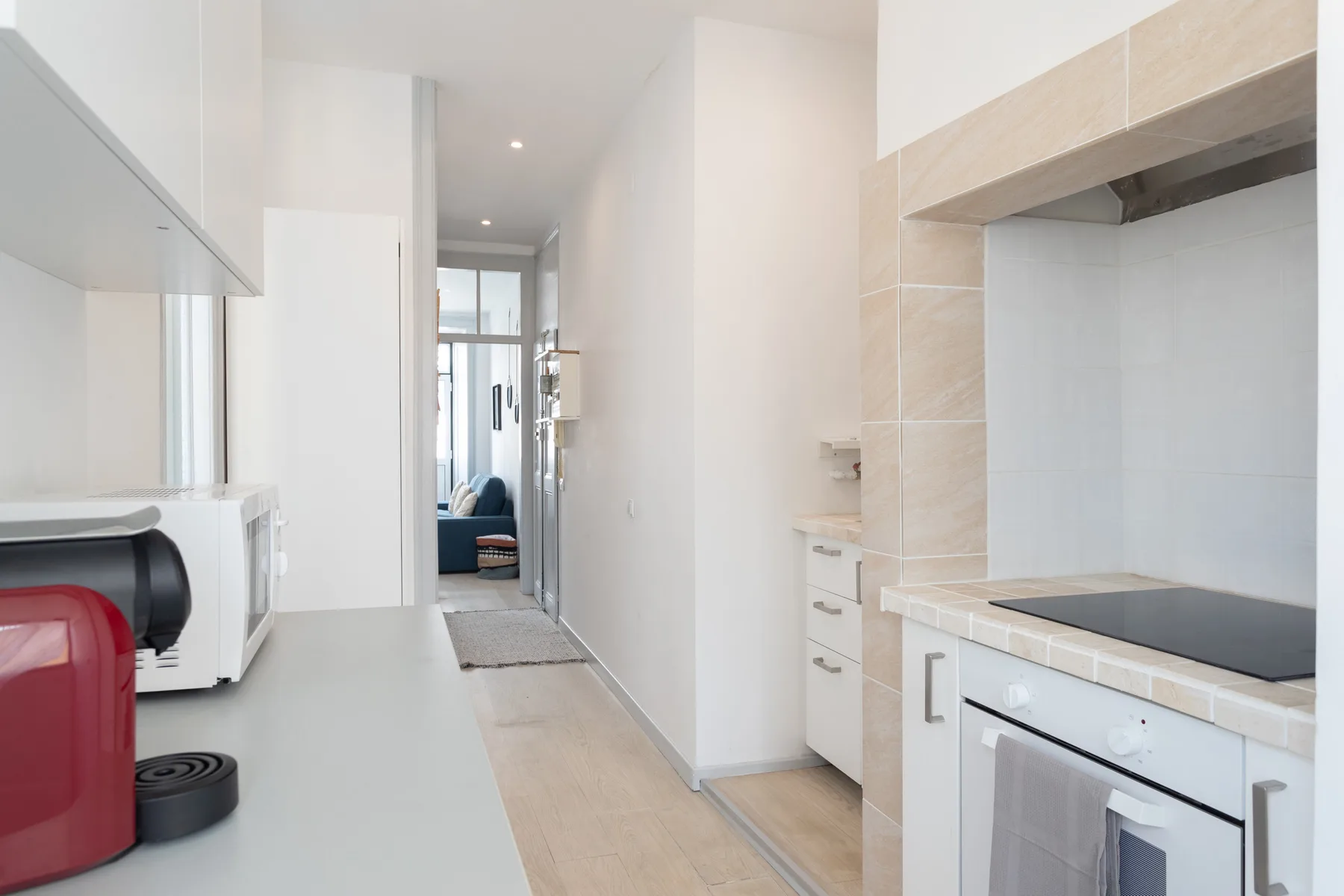 A bright kitchen opens onto a narrow hallway with a living area beyond.