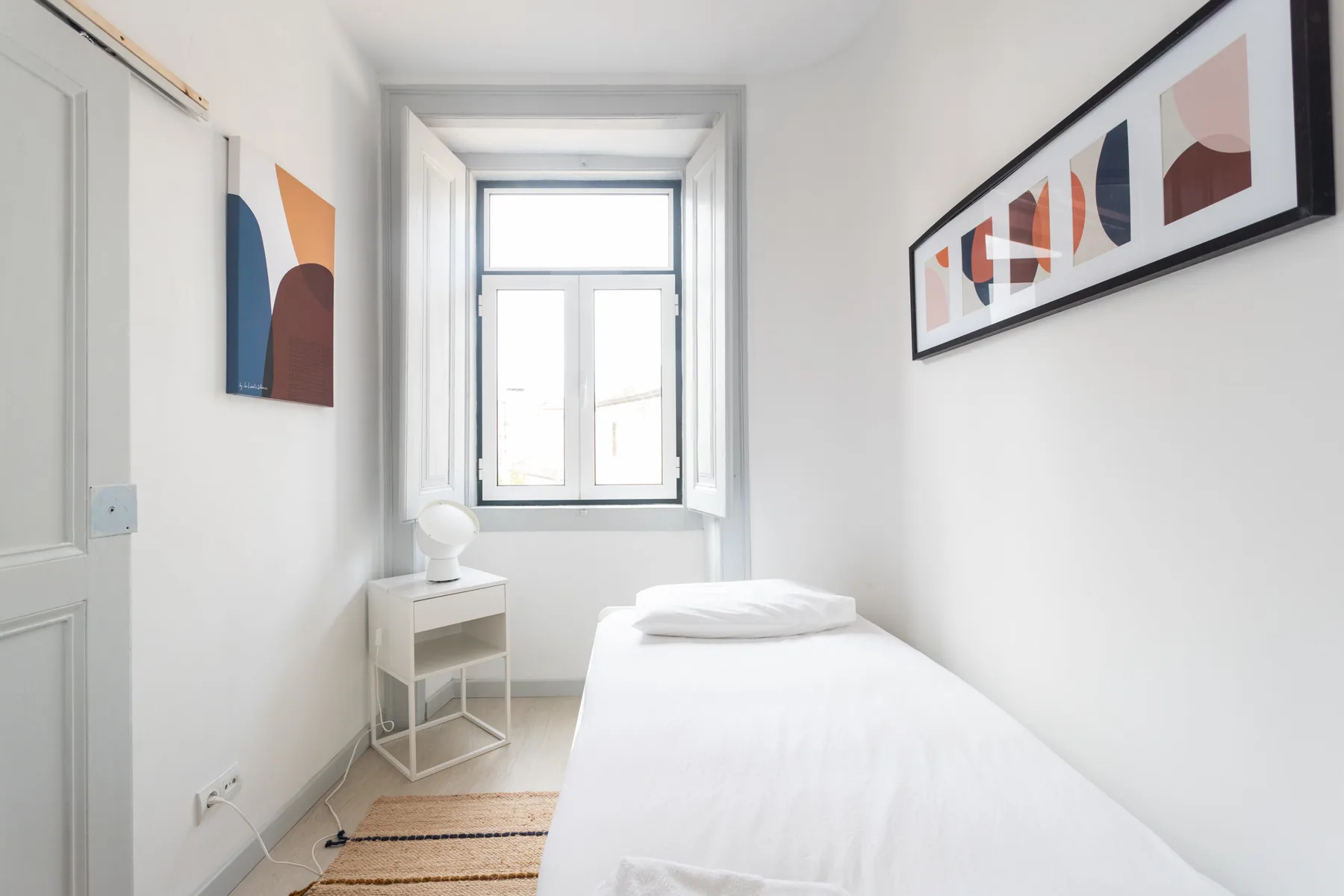 A bright bedroom with a single bed, a window, and framed abstract artwork.