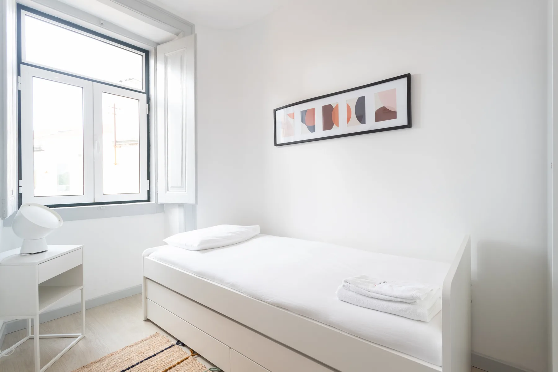 A bright bedroom with a single bed, a large window, and framed wall art.