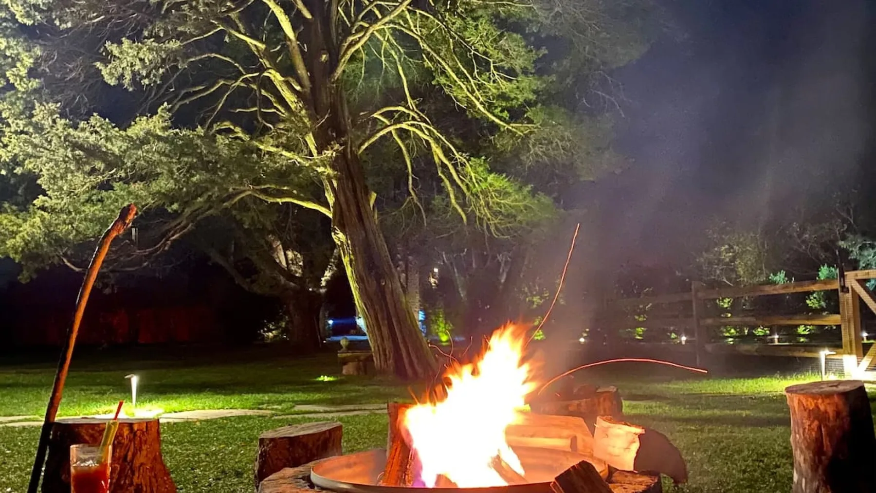 A nighttime garden scene with a bright fire burning among trees and wooden seating.