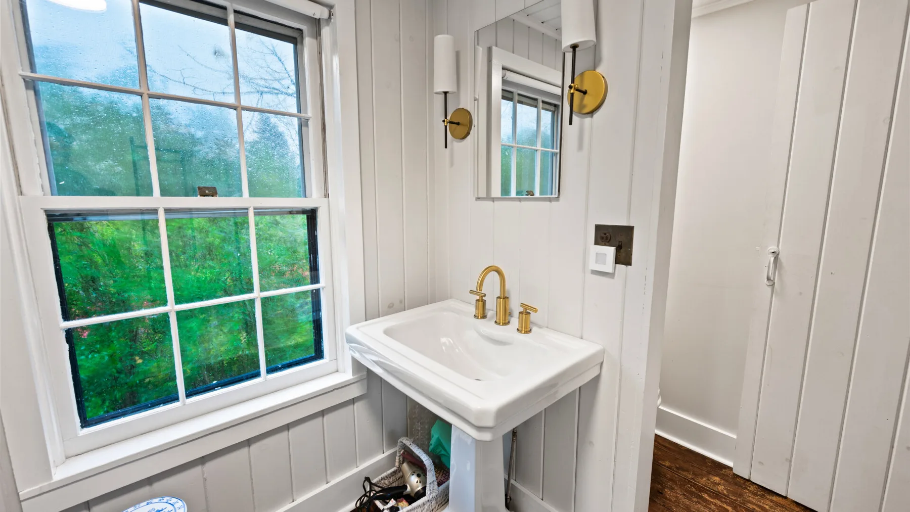 A bathroom vanity sits beside a large window overlooking green trees.