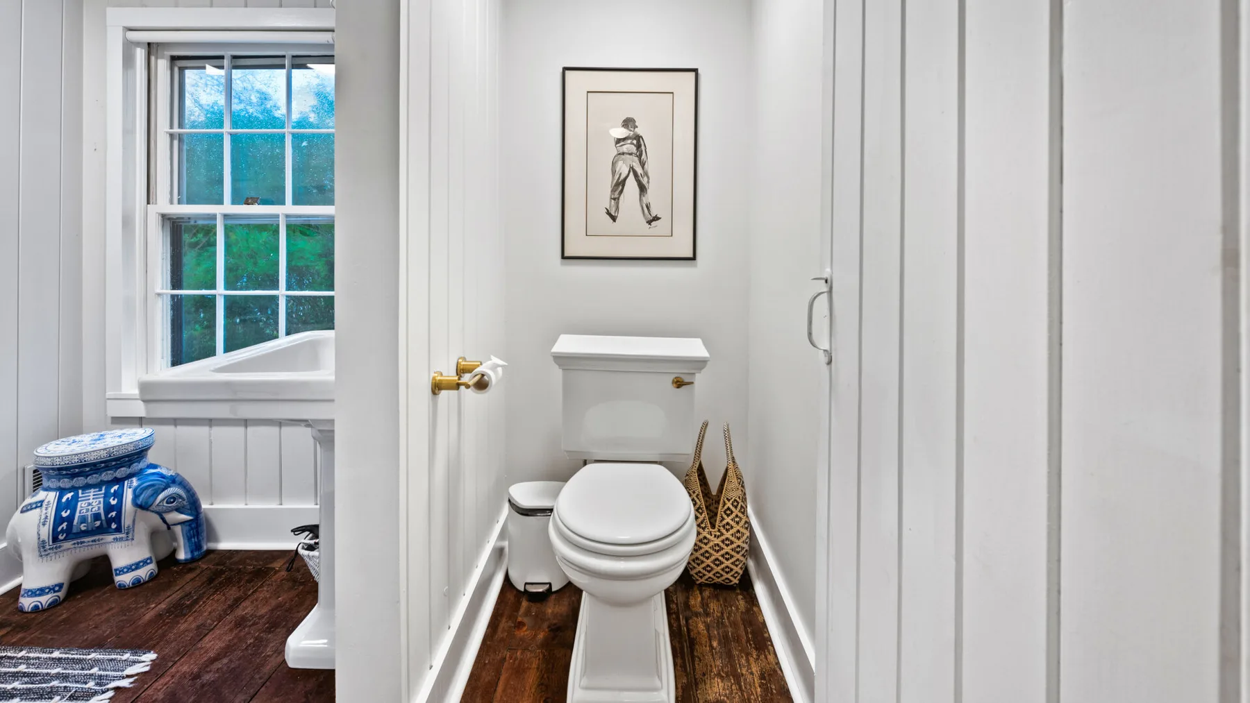 A narrow bathroom with a toilet, framed artwork, and a window.