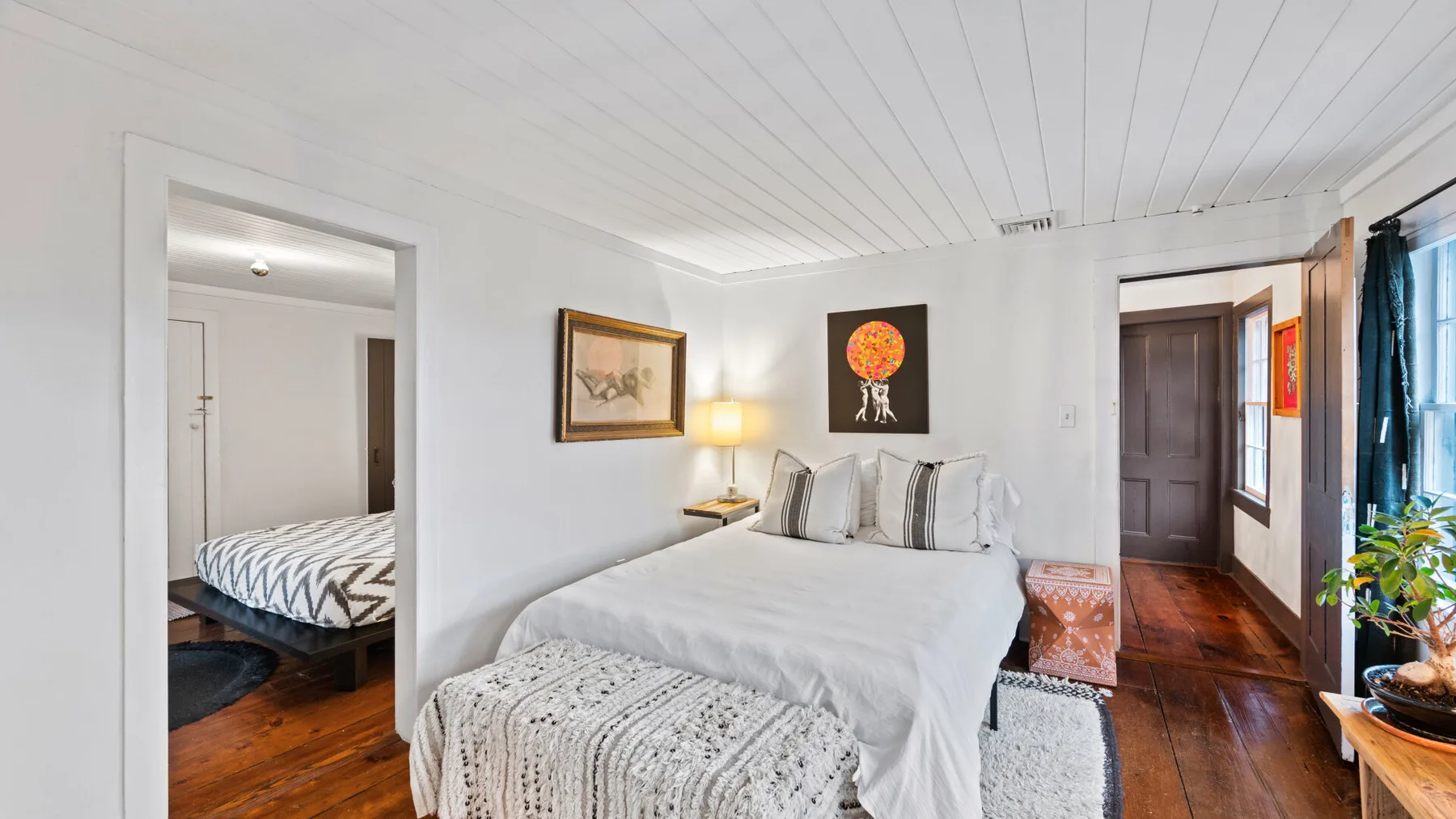 A bright bedroom with a neatly made bed, framed artwork, and wooden flooring.