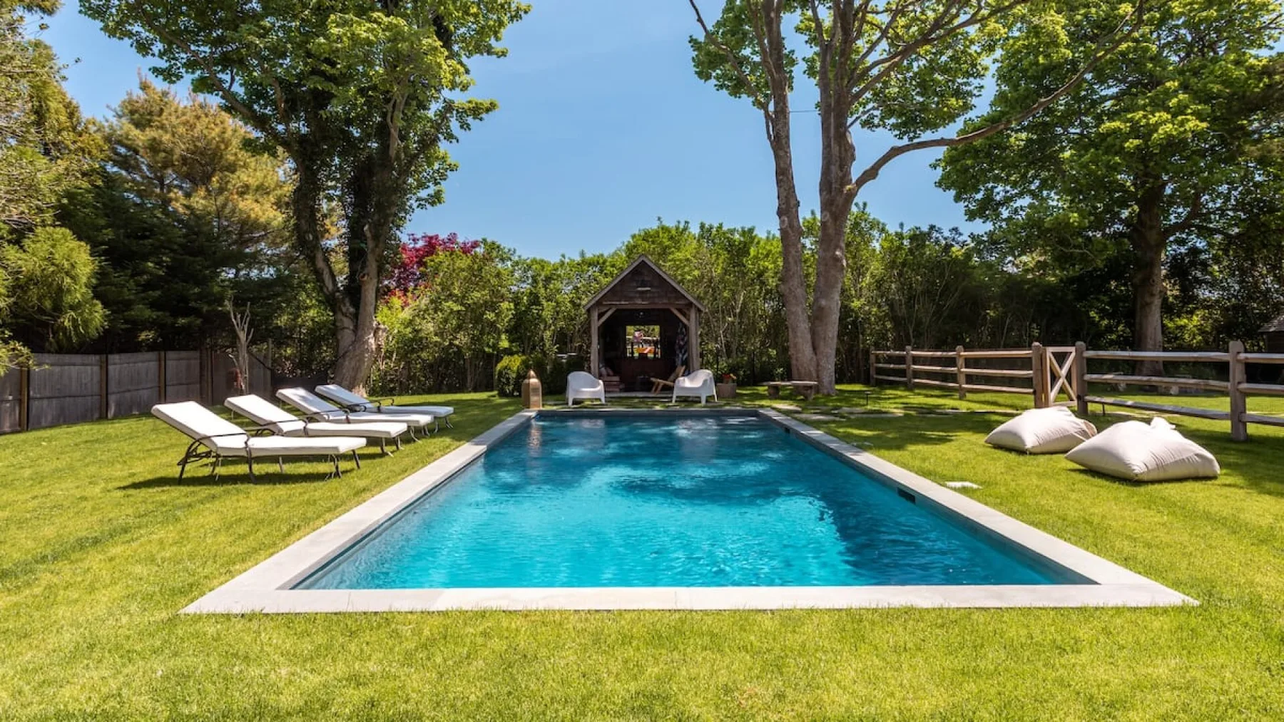 Una piscina al aire libre está rodeada de césped, árboles y asientos para descansar.