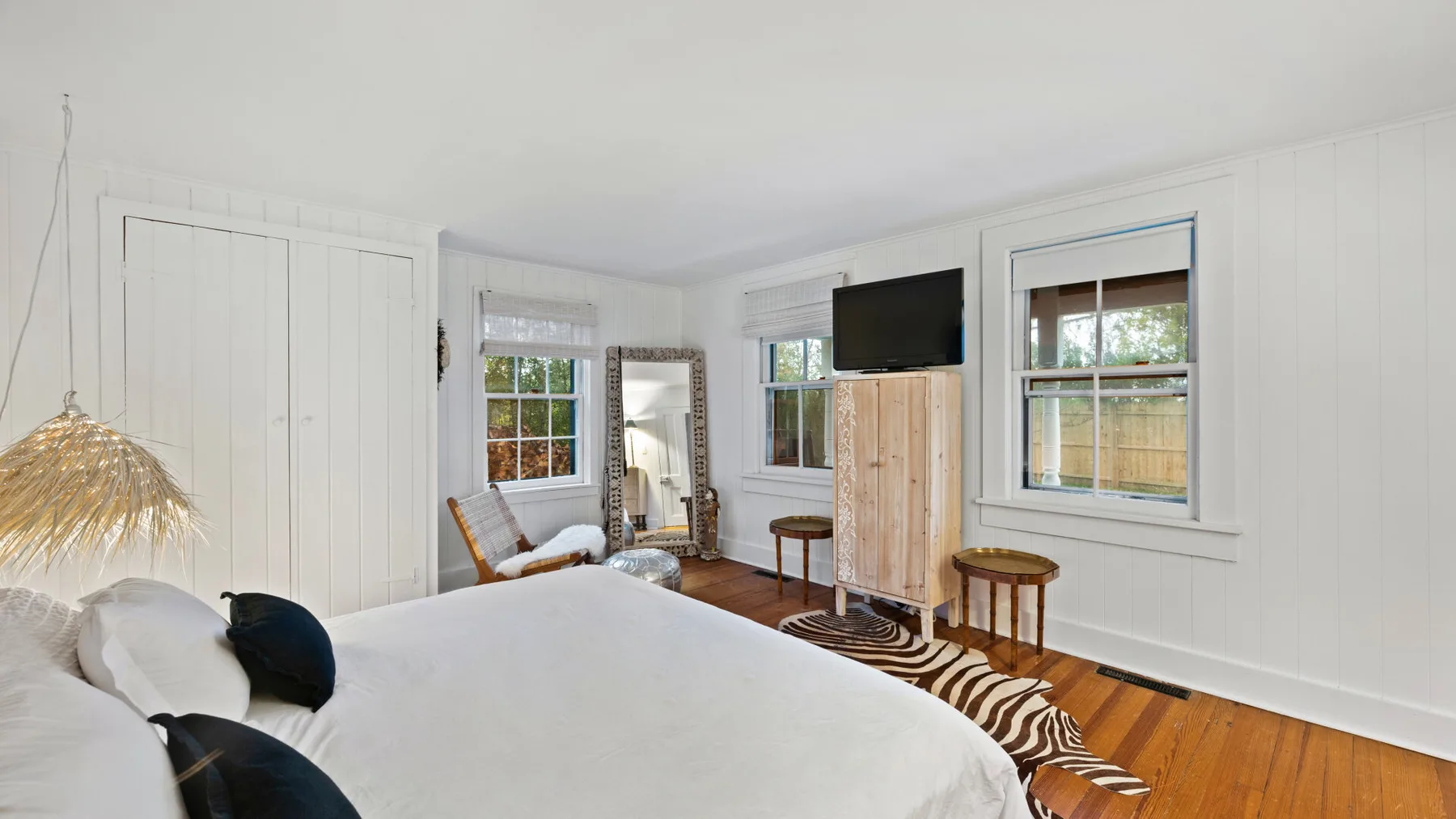 A bright bedroom with a large bed, white paneled walls, and several windows.