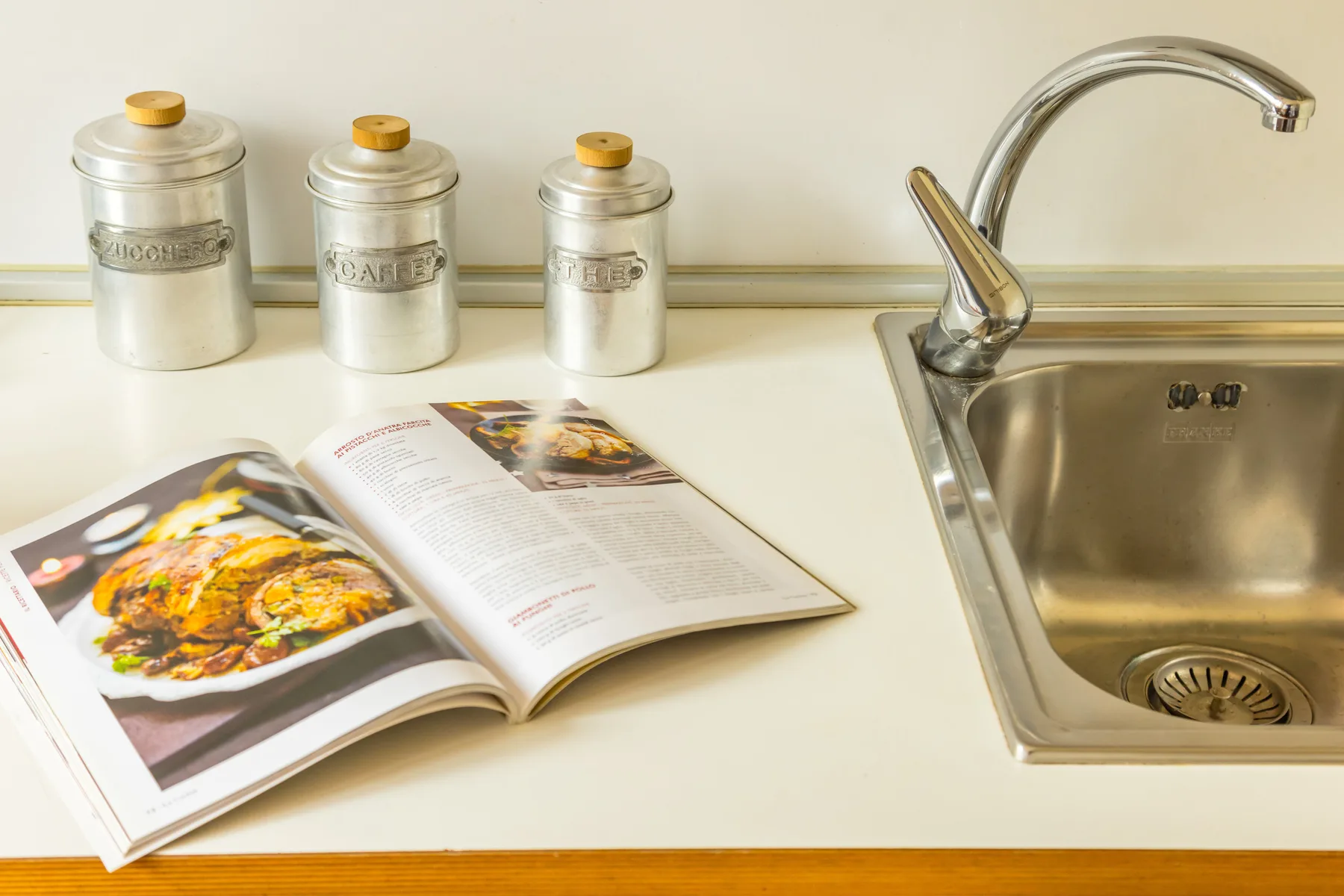 A kitchen countertop with an open cookbook beside a sink and canisters.