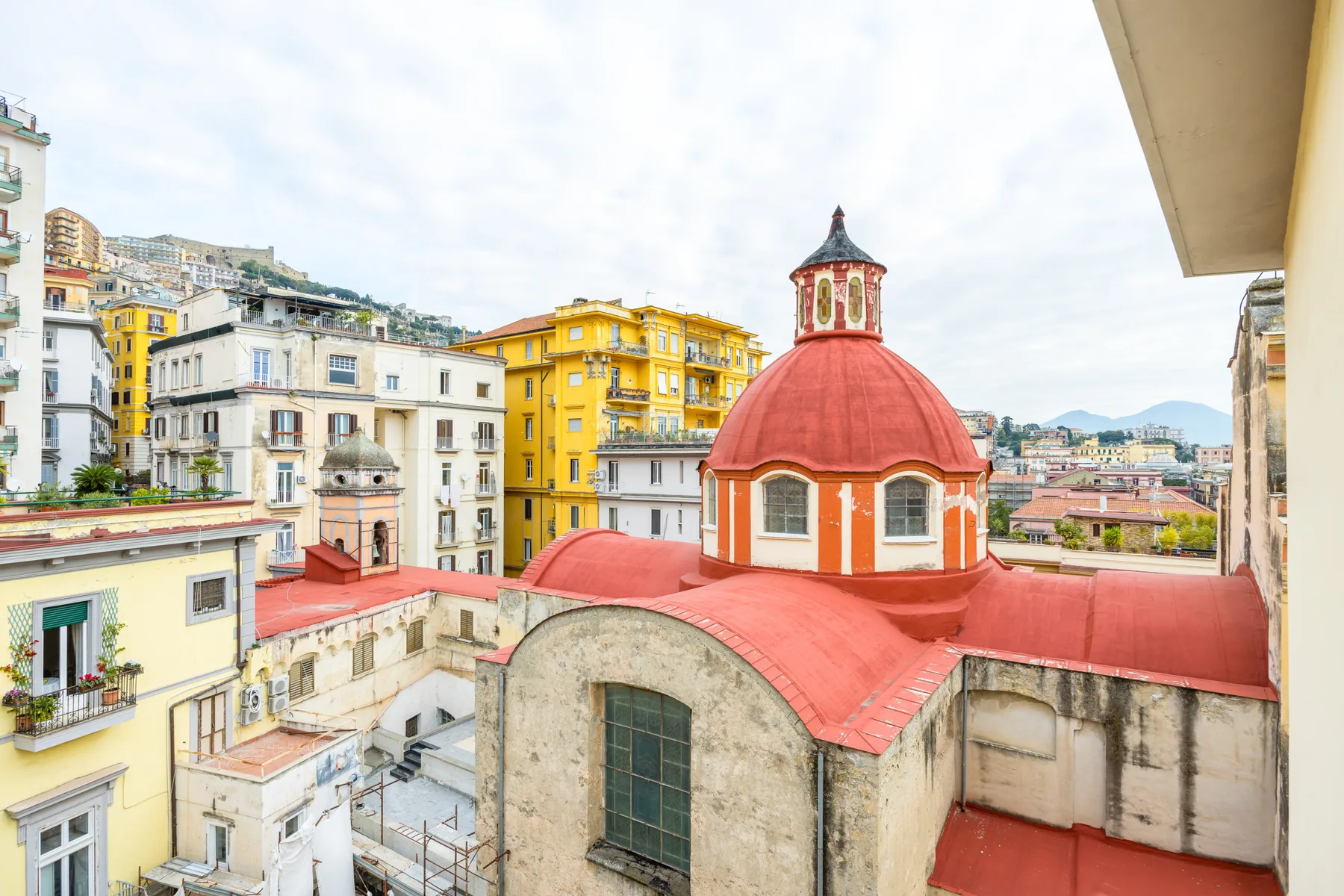 A red-domed church roof stands among colorful city buildings beneath a cloudy sky.