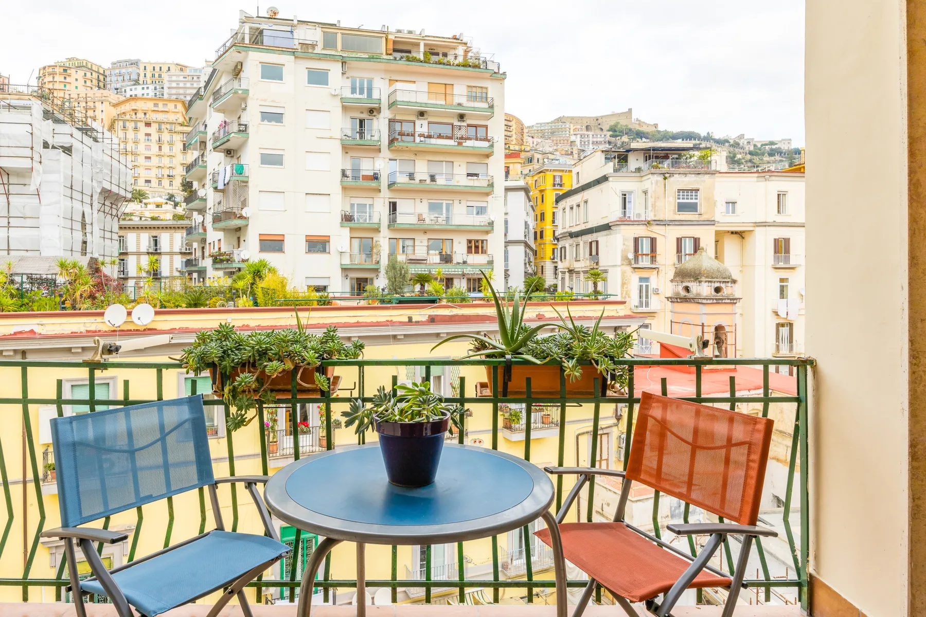 A balcony seating area overlooks nearby apartment buildings, with a potted plant on the table.
