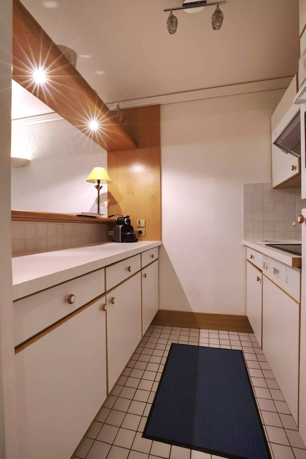 A narrow kitchen with white cabinets, tiled flooring, and a dark floor mat.