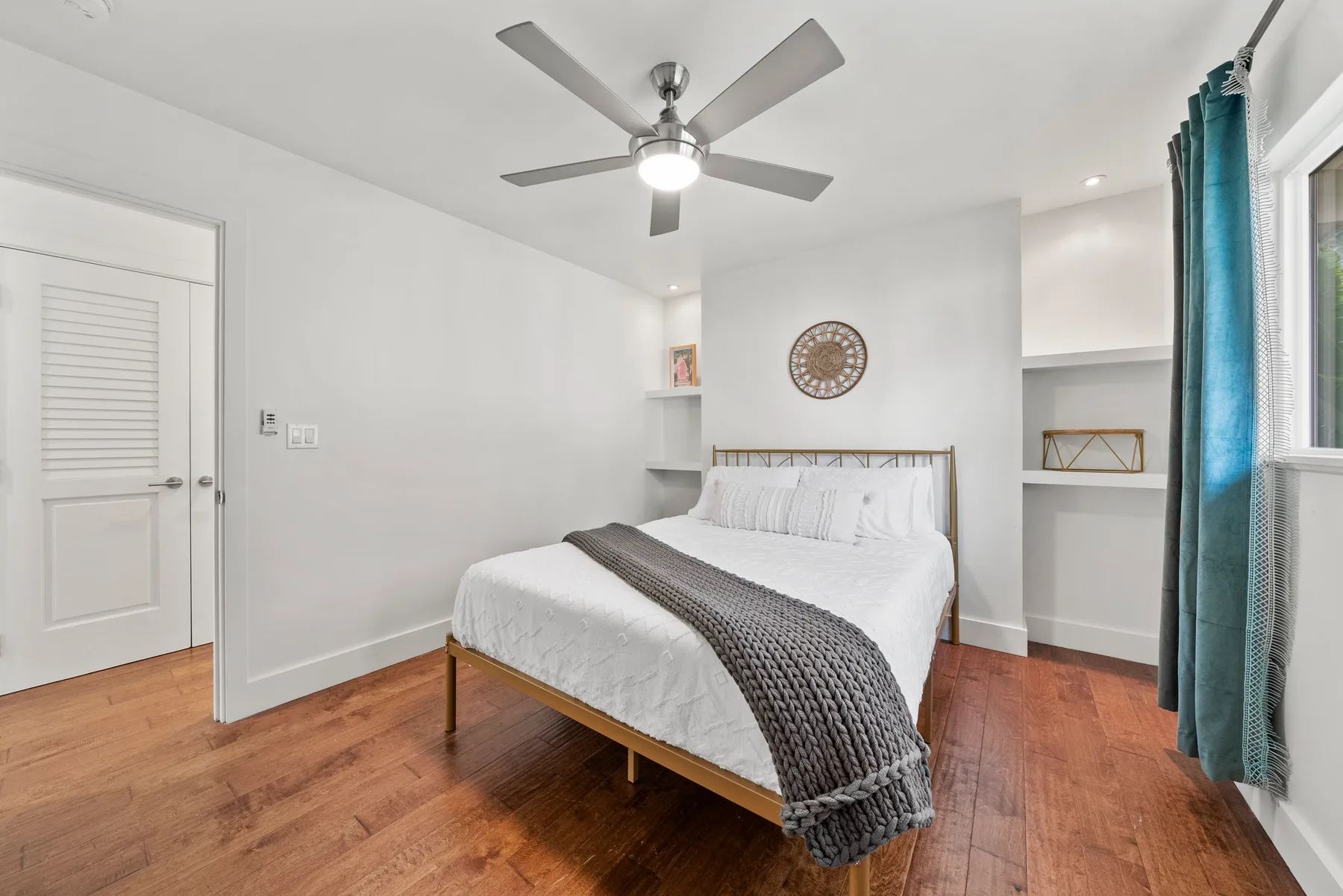 Un dormitorio luminoso con una cama, ropa de cama gris y un ventilador de techo.