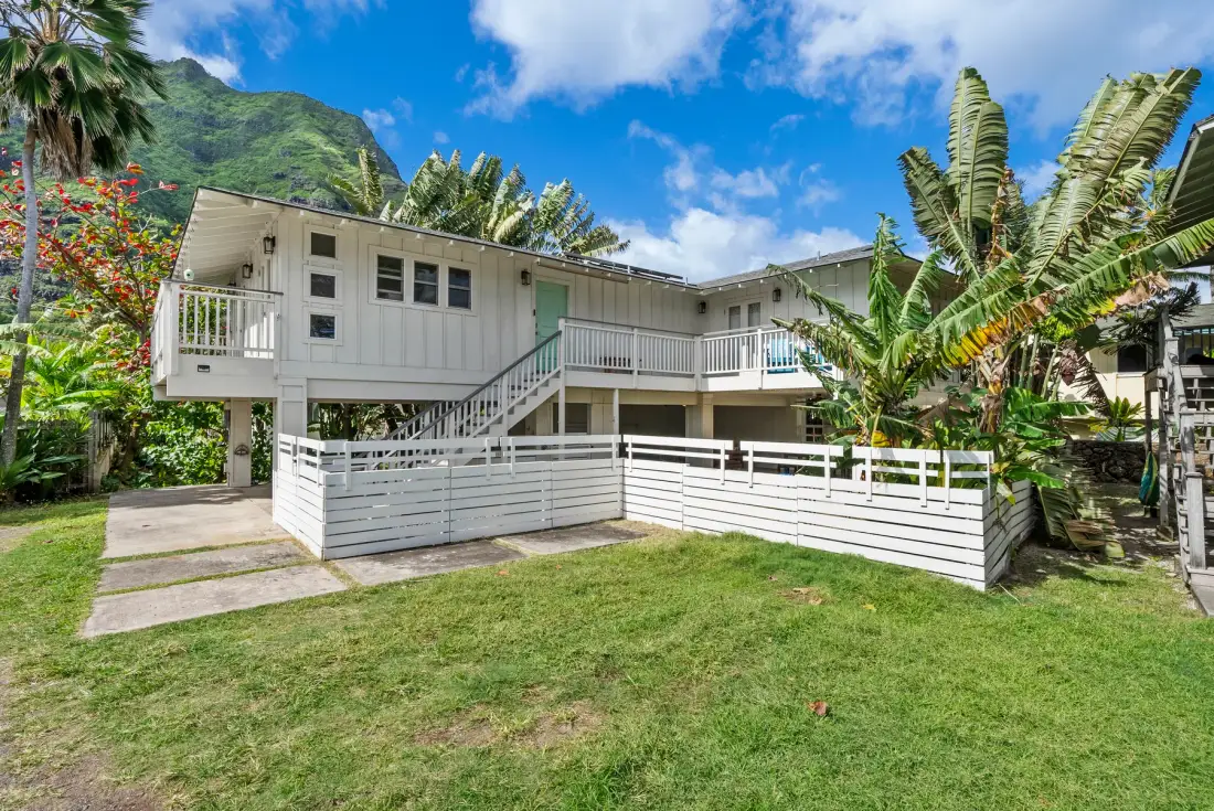 A raised white house stands in a grassy yard surrounded by tropical plants.