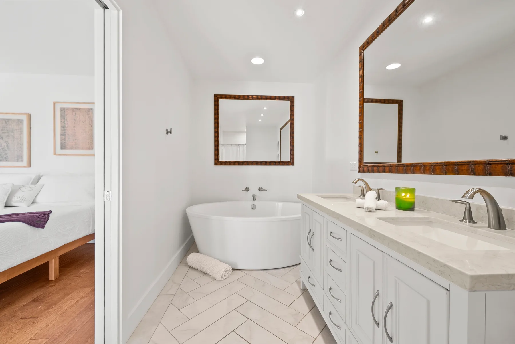 Un baño luminoso con bañera, un tocador largo y un espejo junto a la entrada abierta de un dormitorio.