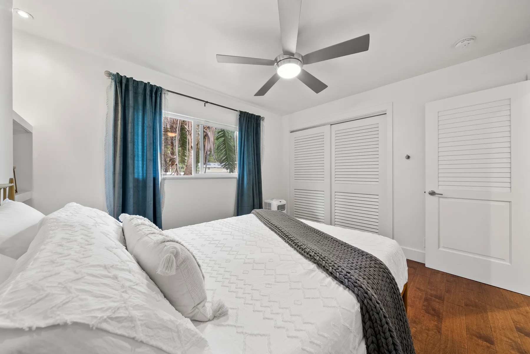 Un dormitorio luminoso con una cama, cortinas azules y puertas blancas con celosías.