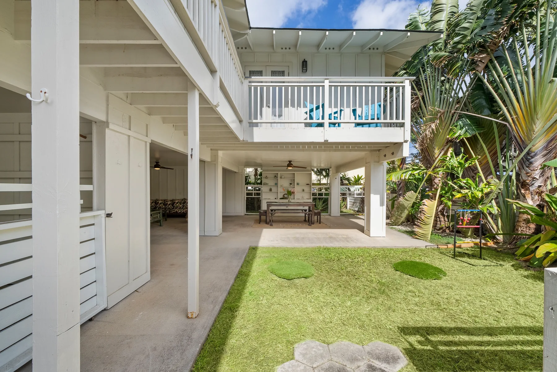 Una casa elevada da a un patio con césped y una zona de comedor sombreada bajo el balcón.