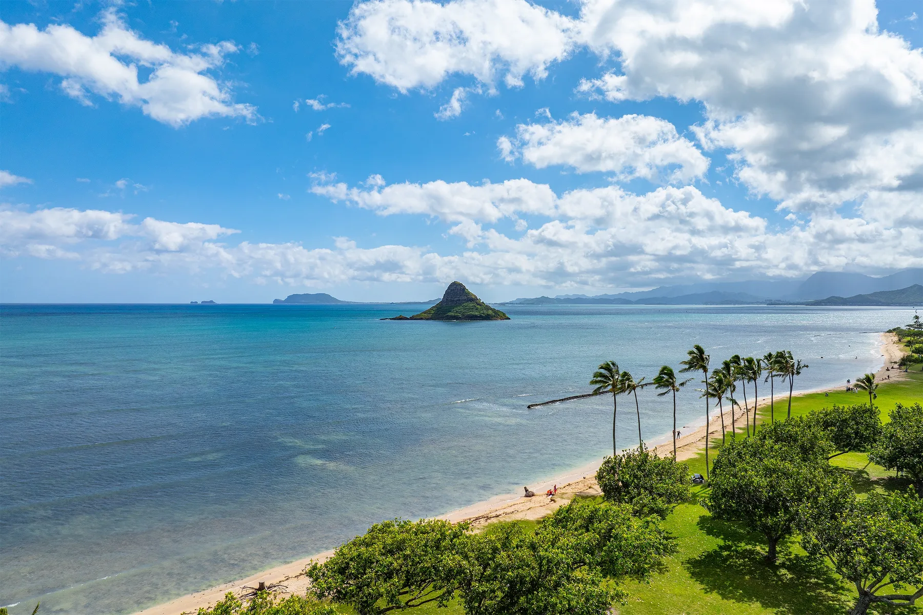 A tropical beach curves along calm blue water beneath a partly cloudy sky.