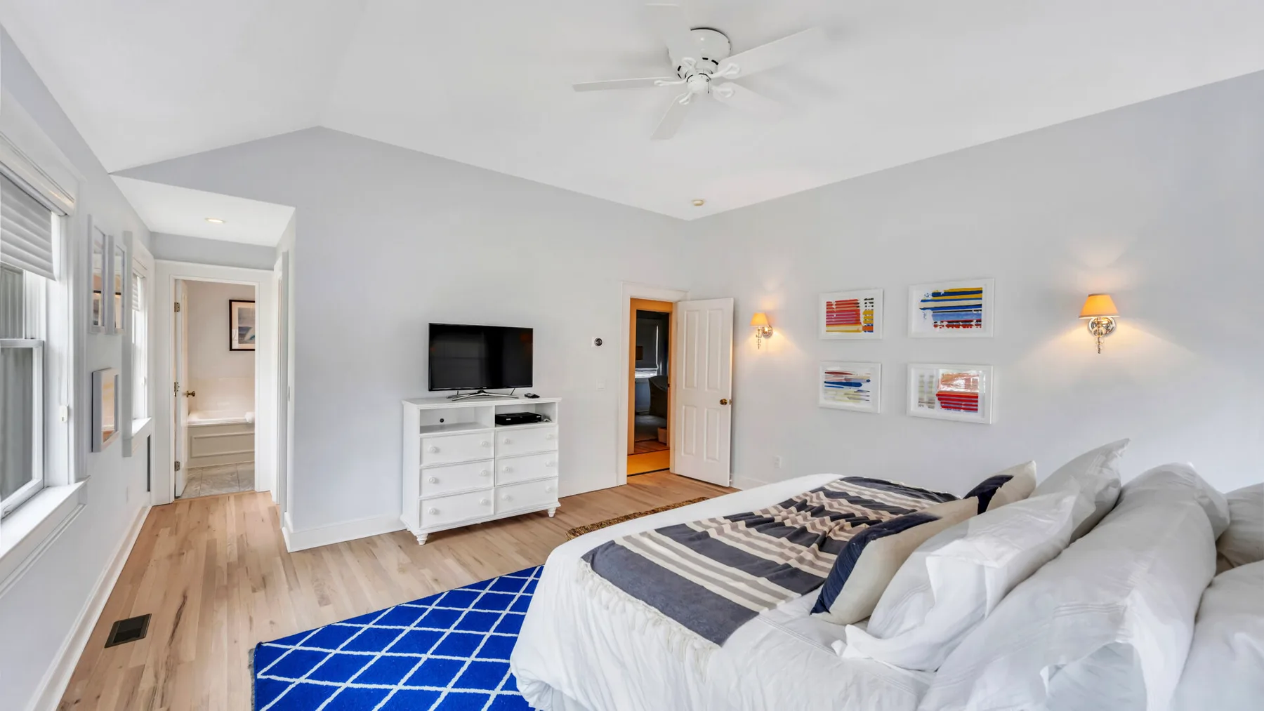 A bright bedroom with white bedding, a dresser, and a blue patterned rug.