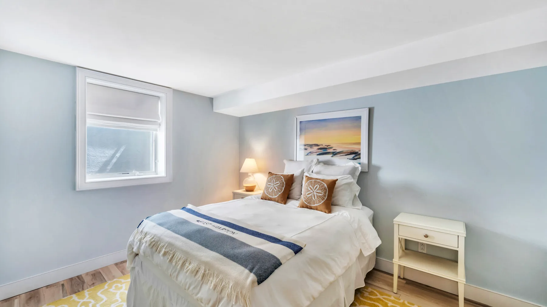 A bright bedroom with a neatly made bed and a framed beach picture.