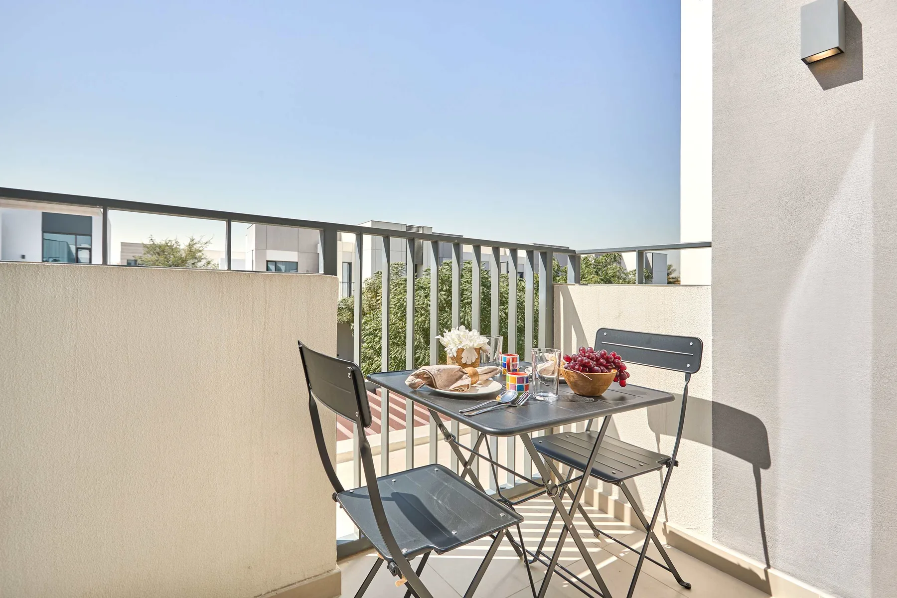 Un balcón soleado con una mesa pequeña y sillas junto a una barandilla metálica.
