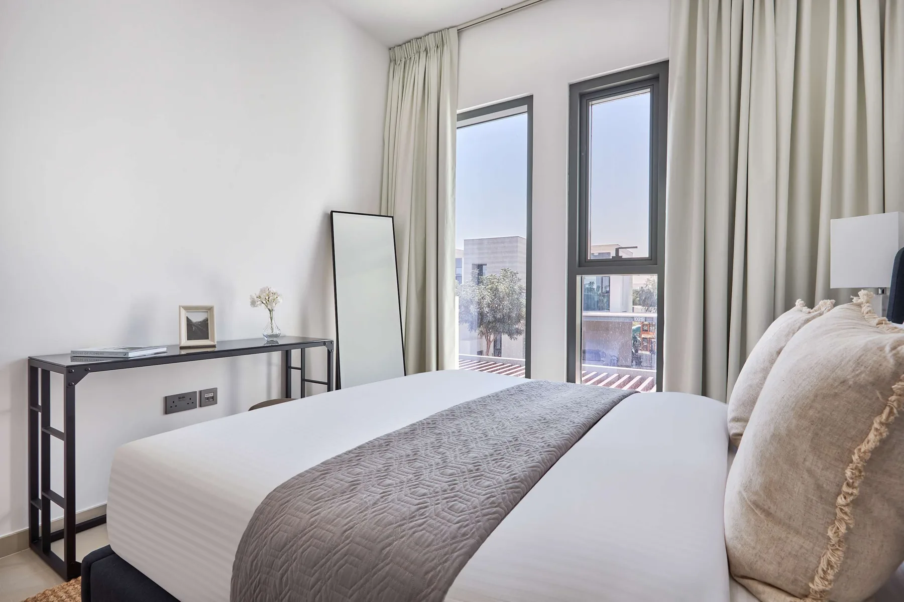 Un dormitorio con una cama bien tendida y ventanas altas junto a cortinas claras.