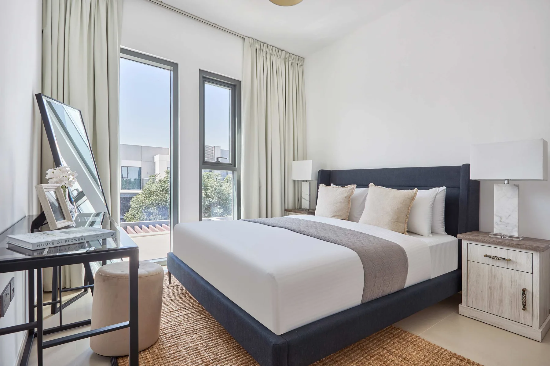 Un dormitorio luminoso con una cama bien tendida junto a ventanas altas y cortinas claras.