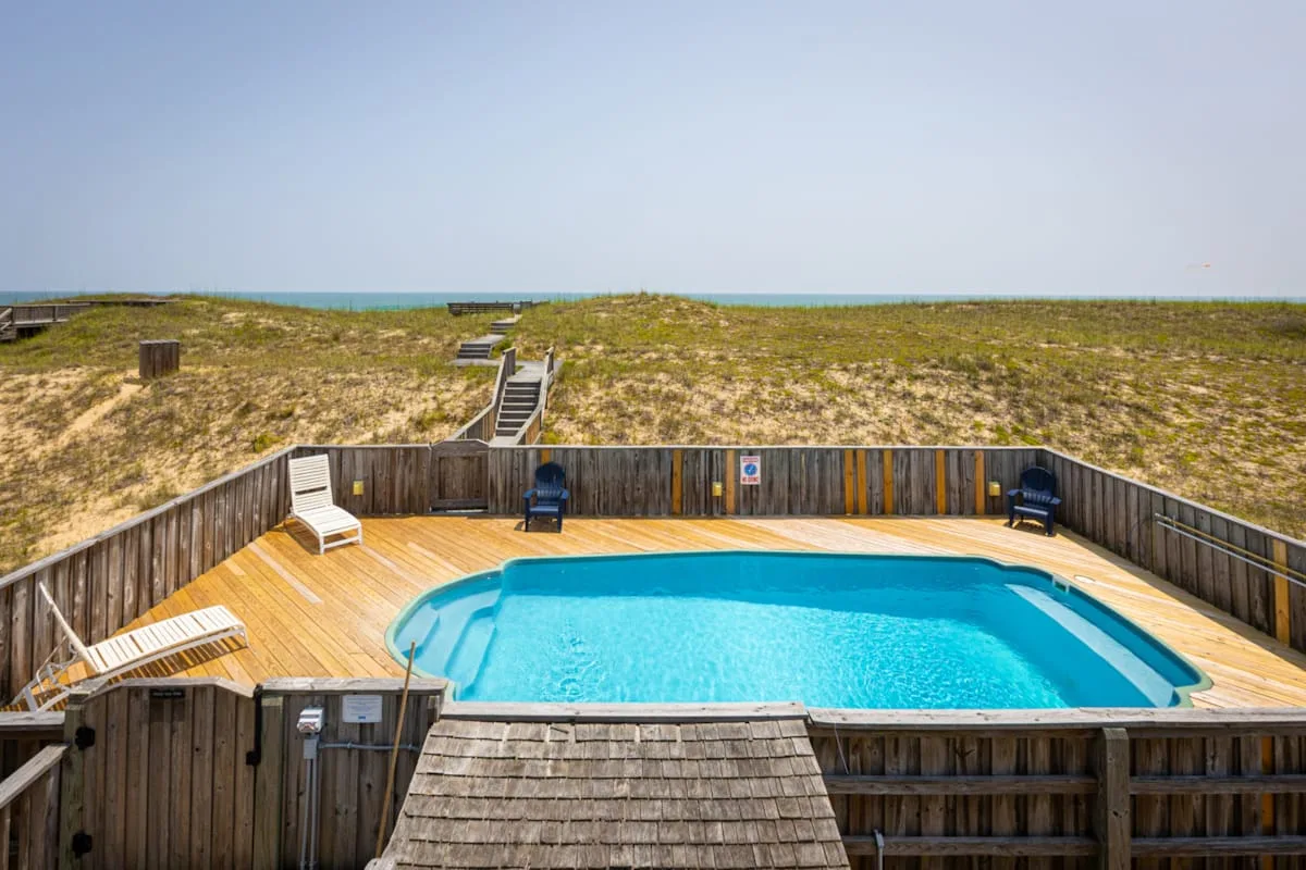 Una piscina al aire libre ocupa una terraza de madera junto a dunas cubiertas de hierba.