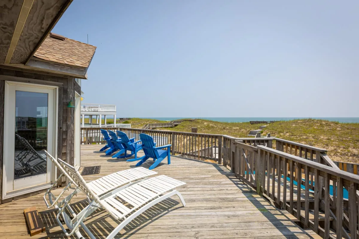 Una terraza de madera junto al mar, con sillas azules y vistas a dunas y al océano.