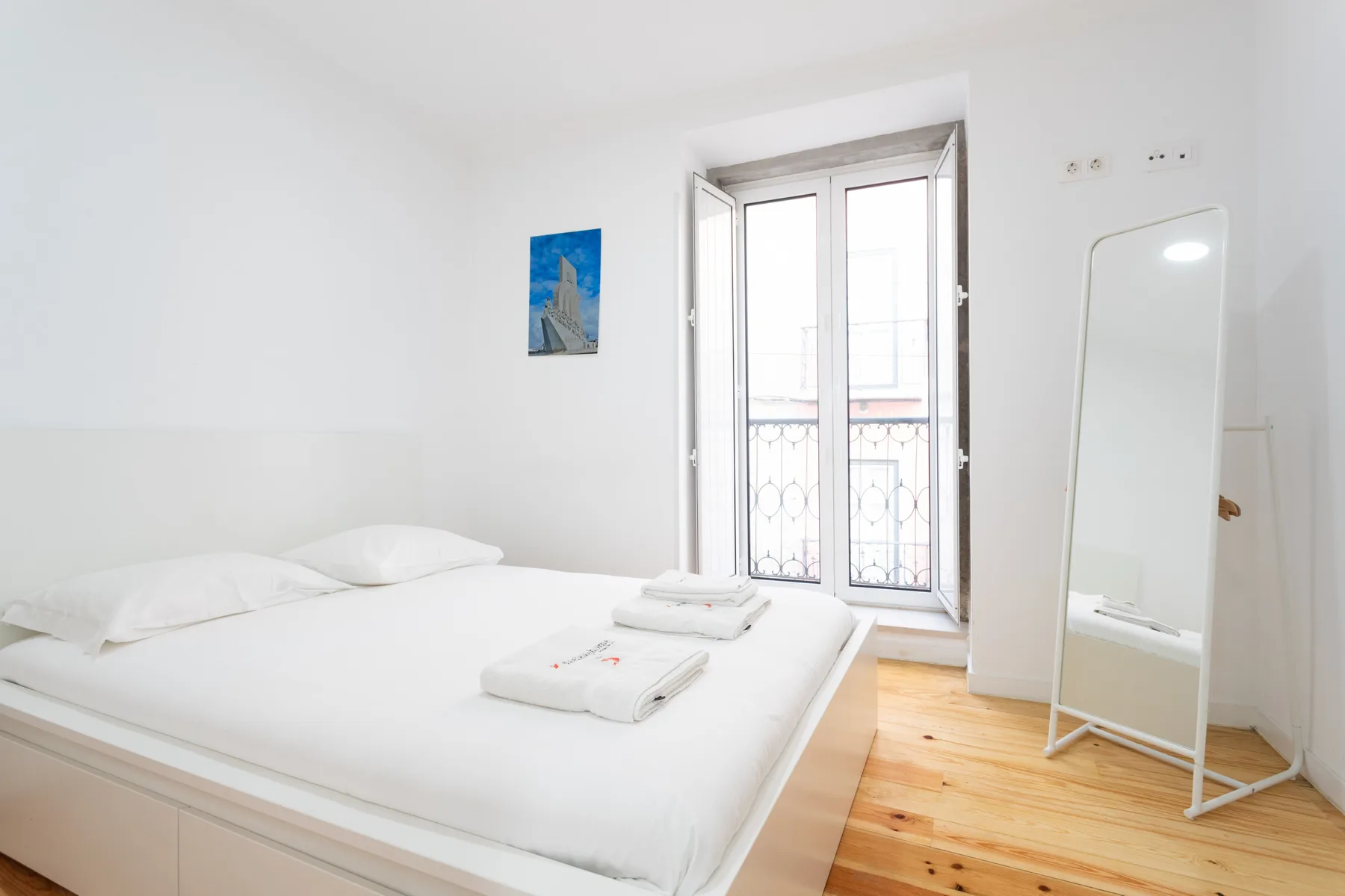Un dormitorio luminoso con ropa de cama blanca, un espejo alto y una puerta abierta al balcón.