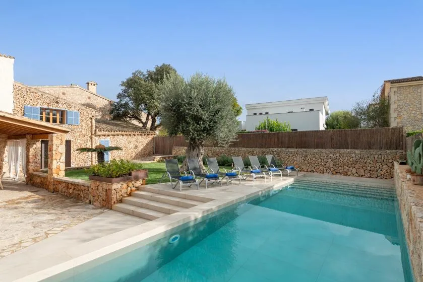 Una piscina con tumbonas está junto a una casa de piedra y un jardín.