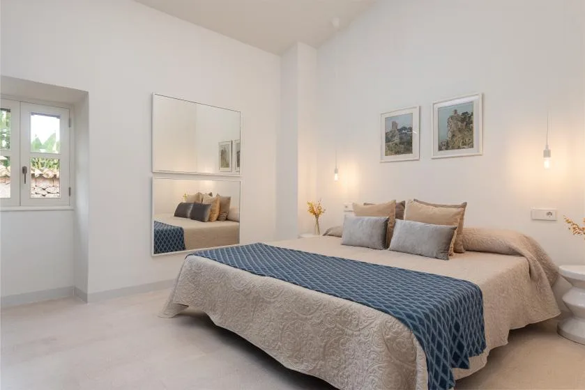 Un dormitorio luminoso con una cama vestida con ropa de cama neutra y una colcha azul estampada.