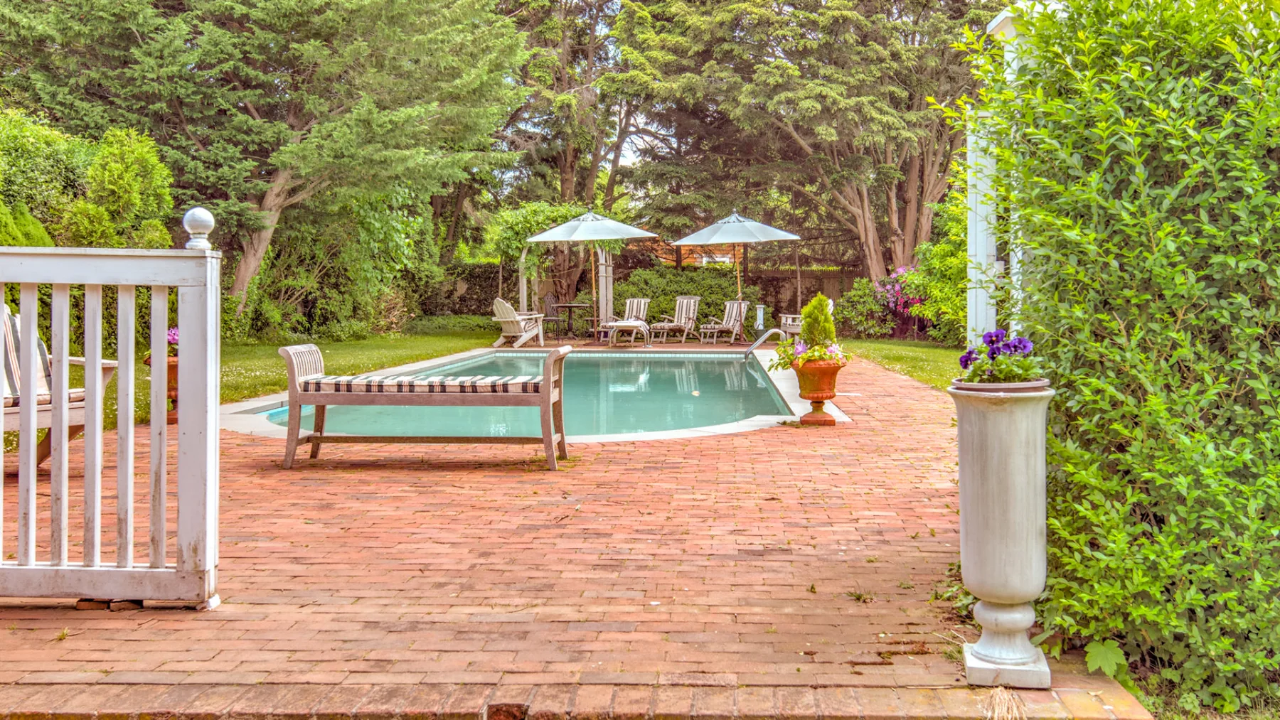 An outdoor swimming pool sits beside a brick patio with seating and umbrellas.