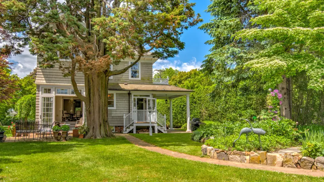 A two-story house sits among trees beside a garden and green lawn.