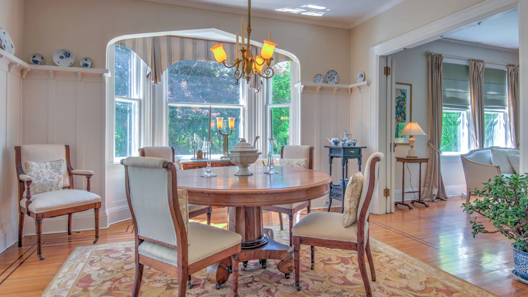 A dining area with a round wooden table and large windows overlooking greenery.
