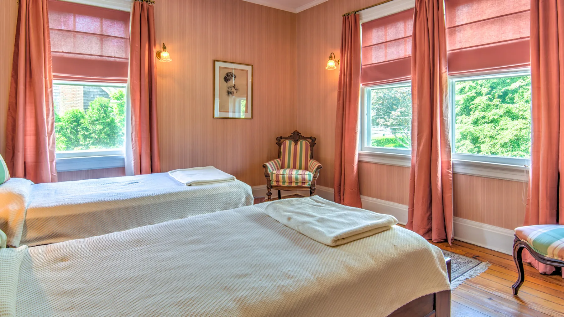 A bedroom with two beds, a small chair, and windows framed by curtains.