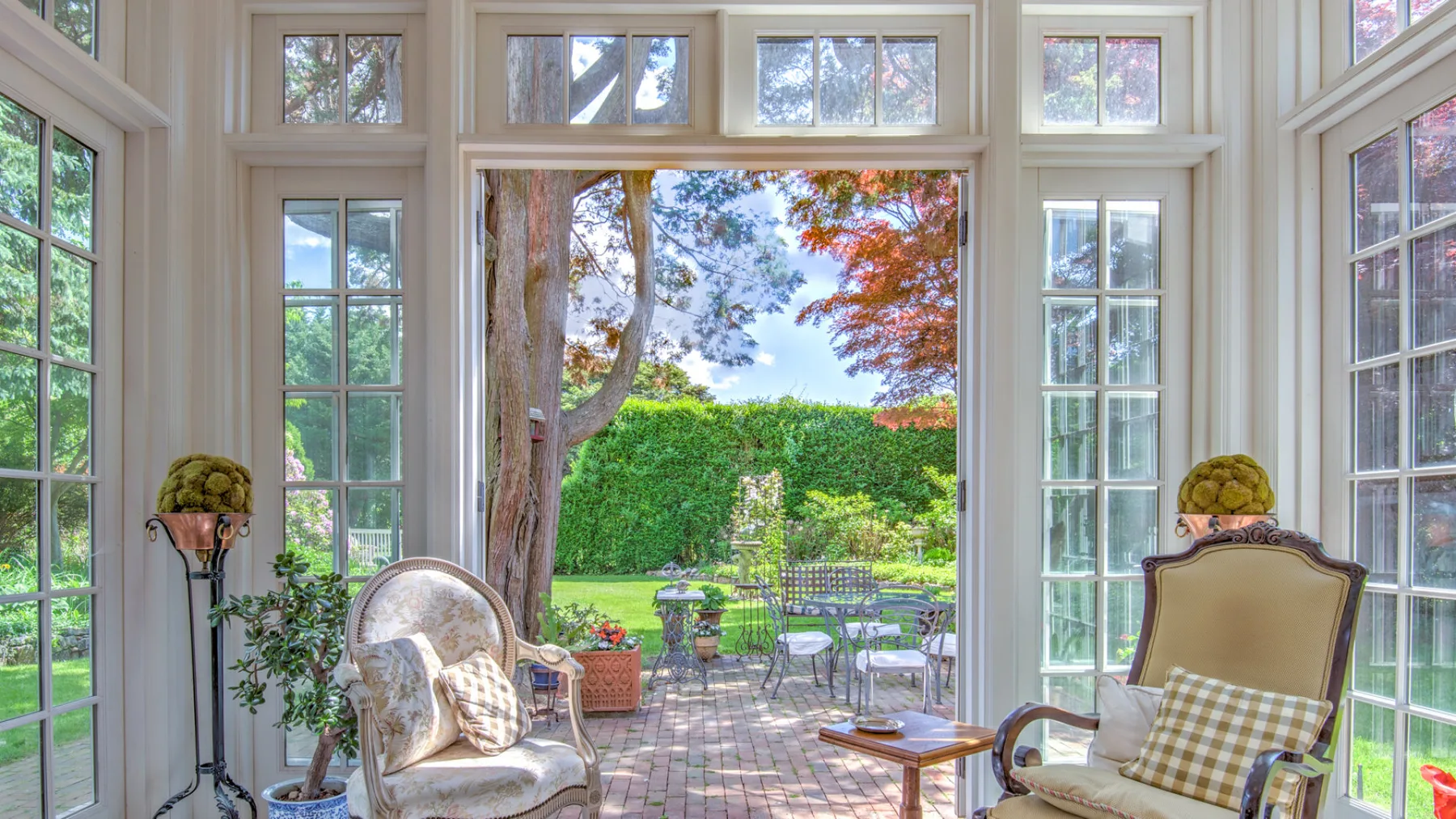 A bright sunroom opens onto a garden patio, with seating beside tall windows.