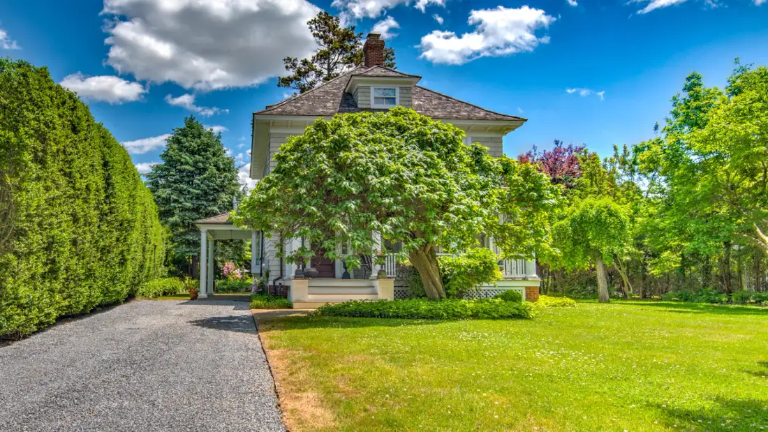 A house stands beside a broad lawn, surrounded by mature trees and shrubs.