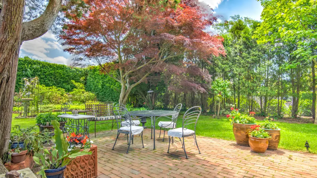 A garden patio with metal seating beneath trees and surrounded by green plants.